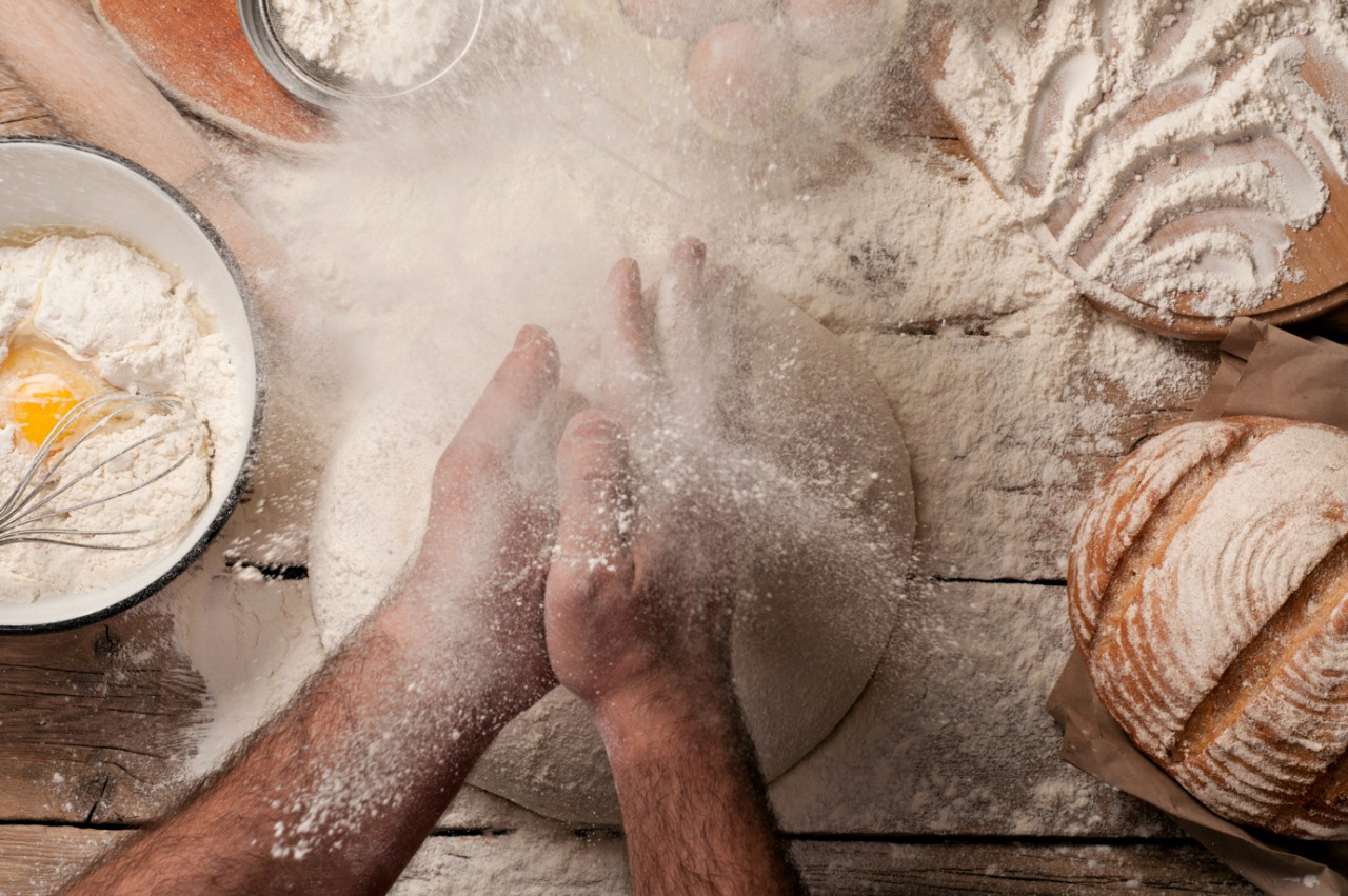 Handwerklich backen bei Poensgen Bemehlte Unterarme und Hände eines Menschen über einem Teig, daneben weitere Backzutaten, Utensilien und ein Laib Brot